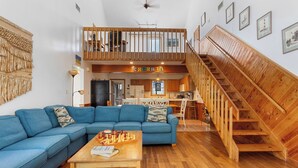 Un salón amplio con un sofá azul, una mesa de centro de madera con un tablero de ajedrez y una escalera de madera que conduce a un nivel superior.