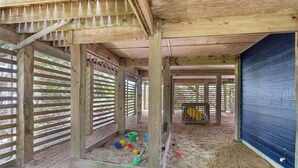 Un espacio al aire libre techado con vigas de madera y una pared azul, que incluye un arenero con juguetes infantiles.