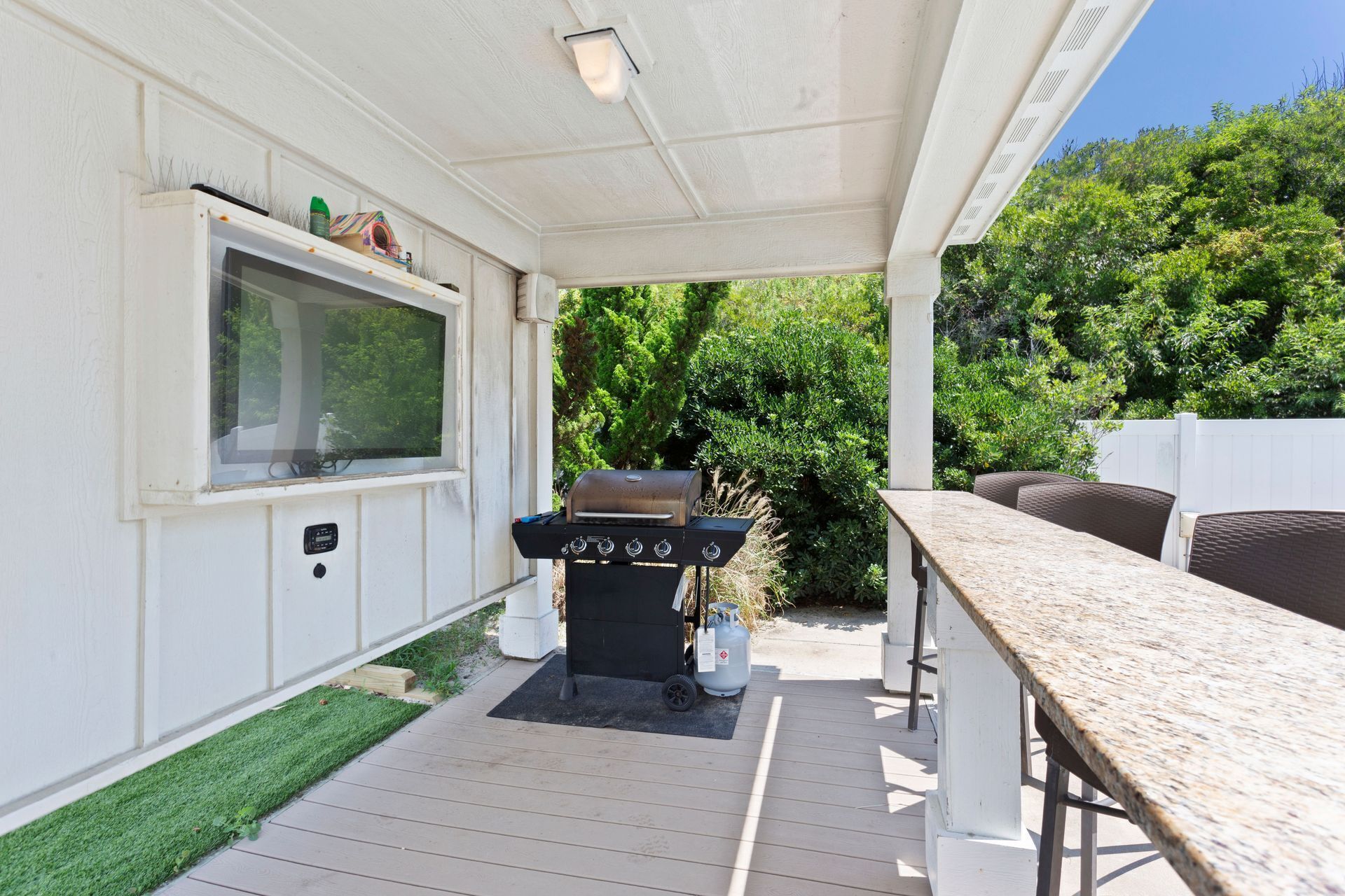 Un patio techado al aire libre con una parrilla, un televisor montado en la pared y una zona de estar con mesa y sillas.