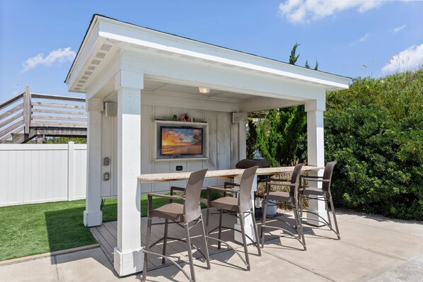 Une terrasse extérieure couverte, équipée d’une table et de quatre chaises, d’un téléviseur fixé au mur et d’un barbecue.