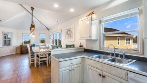 A spacious kitchen with a large window, wooden cabinets, and a dining area with a table and chairs. - The Fifth Line OBX Retreat w/ Pool + Walk to Beach (Corolla)