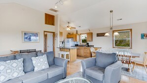 Una sala de estar moderna con un sofá y sillones grises, un comedor con una mesa y sillas de madera, y una cocina con gabinetes de madera y un ventanal.