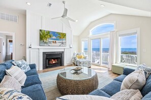 A spacious living room with a fireplace, a large painting of a beach scene, and a balcony with a view of the ocean. - Oceans Eleven: Pool, Hot Tub, Oceanfront (Corolla)