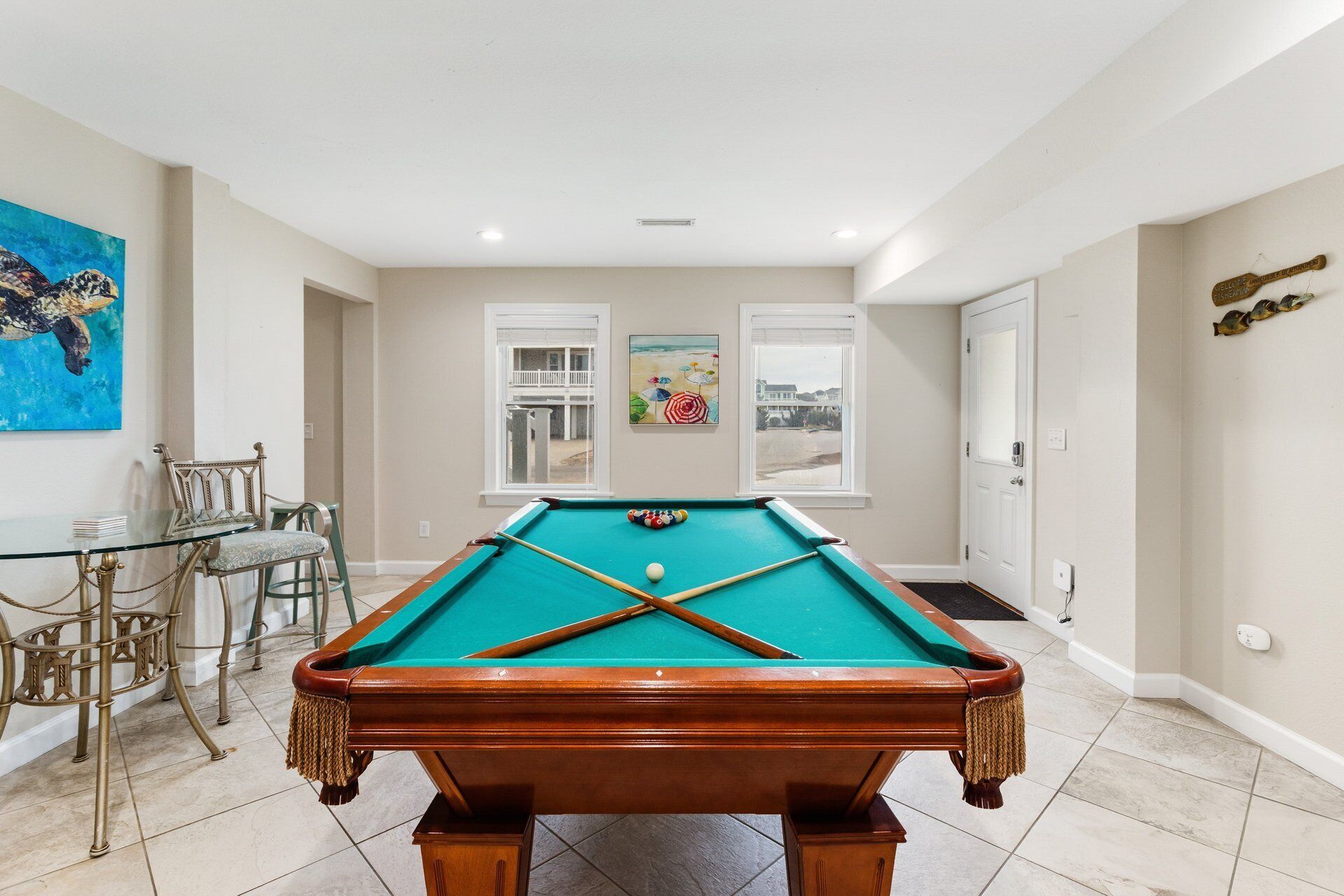 Une table de billard dans une pièce avec une table et des chaises en verre, deux fenêtres donnant sur la plage et un tableau accroché au mur.