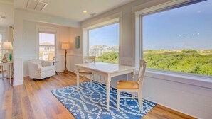 A spacious room with a large window offering a view of the landscape, a white dining table with chairs, a patterned rug, and a cozy seating area with a patterned chair and a lamp. - Ohana Hut: Pool, Hot Tub, 3-Min Walk to Beach (Corolla)