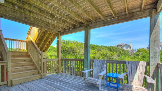 A wooden deck with a staircase leading to a covered area, surrounded by greenery and a house in the background. - Ohana Hut: Pool, Hot Tub, 3-Min Walk to Beach (Corolla)