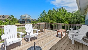 Una terraza de madera con sillas Adirondack blancas y una mesita, con vistas a una zona residencial con casas y árboles.