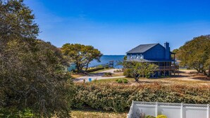 A coastal house with a view of the sea, surrounded by trees and a white fence. - Merryweather: Pool, Hot Tub, Ocean + Sound Access (Duck)