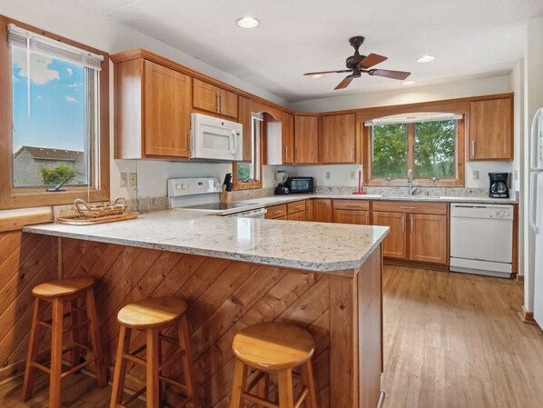 A modern kitchen with wooden cabinets, a large island with stools, a ceiling fan, and a view of the outdoors. - Horizon's Summit: Ocean Views, 2-Min Walk Beach (Duck)