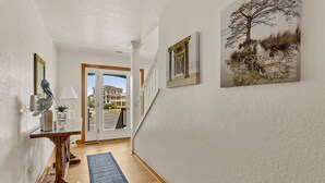 A hallway with a wooden floor, a glass door leading outside, and a staircase with a white railing. - Sol Mates: Pool, Tiki Bar, 3-Min Walk Beach OBX (Duck)