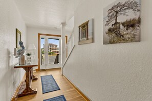 A hallway with a wooden floor, a glass door leading outside, and a staircase with a white railing. - Sol Mates: Pool, Tiki Bar, 3-Min Walk Beach OBX (Duck)