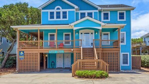 A two-story house with a blue exterior, a front porch with wooden railings, and a small front yard. - Sol Mates: Pool, Tiki Bar, 3-Min Walk Beach OBX (Duck)