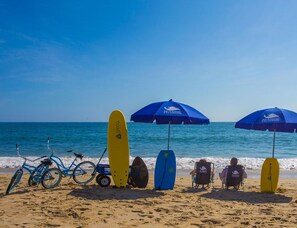 A beach scene with surfboards, bicycles, and beach chairs under umbrellas.