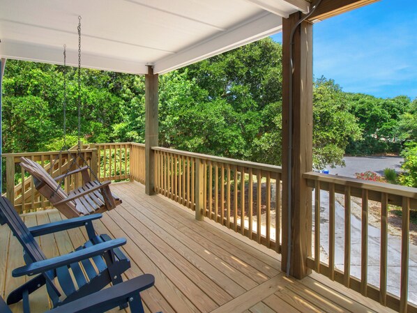 A wooden deck with two Adirondack chairs, a covered area, and a view of a parking lot and trees. - Luna Blu Cottage: Pool, Hot Tub, Duck OBX Retreat (Duck)