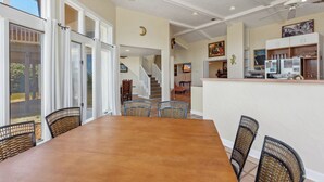 Ruime eethoek met een grote houten tafel en rieten stoelen, een keuken met witte kasten en uitzicht op de omgeving door grote schuifdeuren.