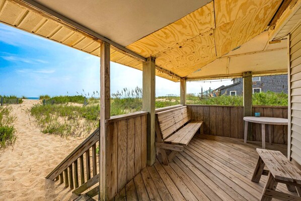 A wooden deck with a bench and table, overlooking a sandy beach and ocean.