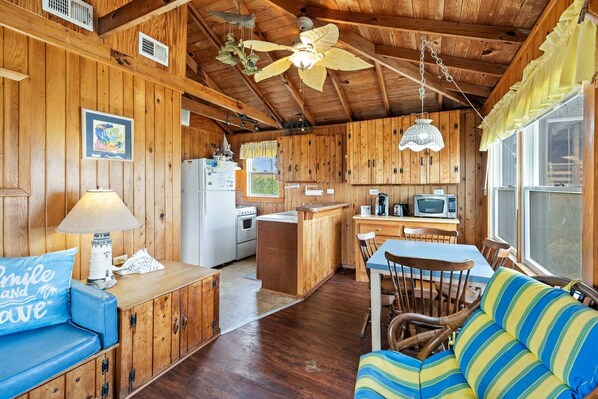 A cabin interior with wooden walls, a kitchenette, a dining area with a table and chairs, and a sofa. - Neaptide: Oceanfront Escape for Two w/ Deck Views (Kitty Hawk)