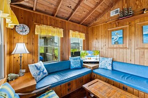 A cabin interior with a wooden ceiling, a clock, a television, and a blue sofa with decorative pillows. - Neaptide: Oceanfront Escape for Two w/ Deck Views (Kitty Hawk)