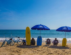 A beach scene with surfboards, bicycles, and beach chairs under umbrellas. - Sea Spray: Steps to Beach w/ Ocean Views OBX (Kitty Hawk)