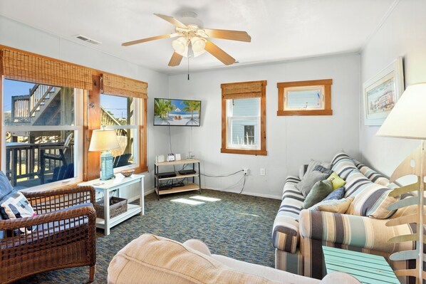 A living room with a ceiling fan, a TV, a wicker chair, a striped sofa, and a view of a wooden deck. - Jo's Bungalow: Walk to Beach, Dog-Friendly OBX (Kitty Hawk)