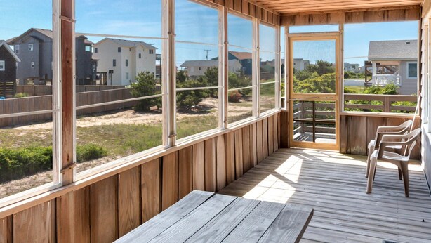 Terraza techada al aire libre con bancos de madera y amplios ventanales que ofrecen vistas a casas vecinas y follaje.