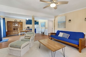 Een woonkamer met een blauwe bank, rieten stoelen, een houten salontafel en een plafondventilator.