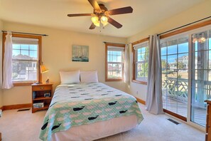 A bedroom with a bed, a ceiling fan, a nightstand, a lamp, and large windows with curtains.
