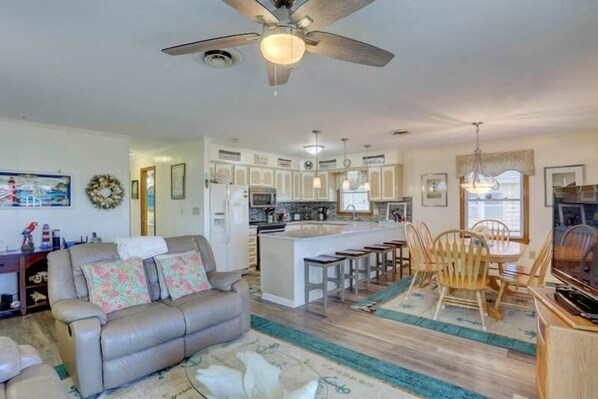 A spacious living room with a sofa, a dining area with wooden chairs, a kitchen with white cabinets, and a ceiling fan.