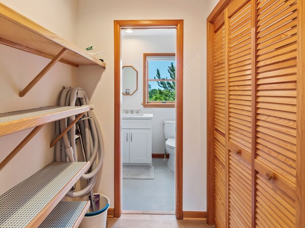 A narrow hallway leading to a bathroom with a toilet, a sink, and a window with a view of greenery. There are shelves with cleaning supplies and a vacuum cleaner. - The Shipley: Soundside Escape Near Beach & Trails (Kitty Hawk)