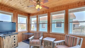 A cozy room with wooden walls and ceiling, a ceiling fan, a TV on a cabinet, a small table, two striped chairs, and large windows offering a view of the neighborhood.