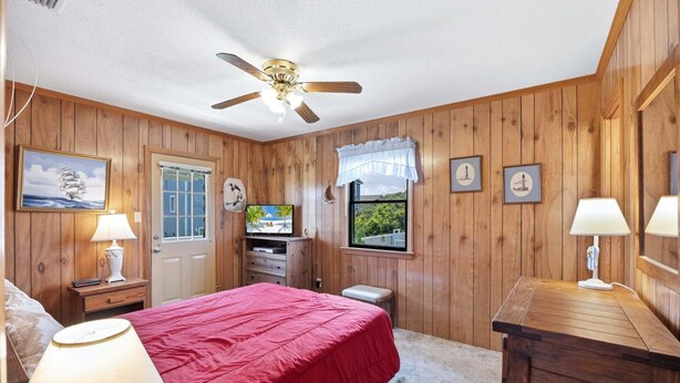 A bedroom with wooden paneling, a bed with a red bedspread, a ceiling fan, a television, and framed pictures on the wall. - 5215 Edwards * 3 Min Walk to Beach (Kill Devil Hills)