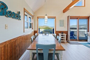 A dining area with a long wooden table, blue chairs, and a view of the beach through large windows. - 5237 Carpe Dune * Oceanfront (Kill Devil Hills)