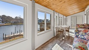 Habitación con amplios ventanales que dan a una masa de agua, techo de madera y sofá blanco con un cojín de colores.