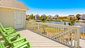 Une terrasse en bois avec des chaises vertes, un bâtiment blanc avec une porte, et des maisons le long d’un plan d’eau.