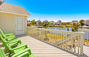 A wooden deck with green chairs, a white building with a door, and houses along a water body.