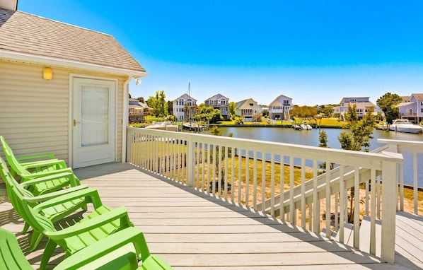 A wooden deck with green chairs, a white building with a door, and houses along a water body.