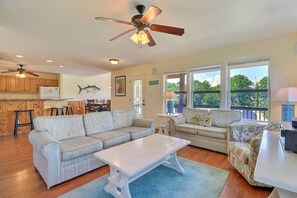 A spacious living room with a large striped sofa, a white coffee table, and a patterned armchair. There is a ceiling fan, a dining area with wooden cabinets, and a view of the outdoors through large windows.