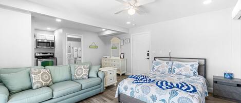 A bedroom with a bed, a sofa, a dresser, a ceiling fan, and a kitchenette in the background.
