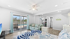 A modern bedroom with a large bed, a ceiling fan, a sliding glass door to a balcony, and a kitchen area visible through an open door. - Sleeps 22, Private Pool, Walk to Beach, Times Square Restaurants! (Fort Myers Beach)