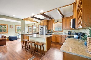 A spacious kitchen with wooden cabinets, a central island, and a dining area with a brown leather couch and wooden stools.