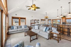 A spacious living room with a large sofa, a coffee table, and a dining area with chairs and a bar. - Ferrari II *SEMI-OCEANFRONT*Pool*HotTub*ArcadeGame (Nags Head)