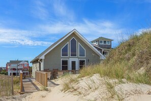 A beachfront house with a prominent gable roof, a wooden deck, and a sandy beach in the foreground. - 6137 Harper * Oceanfront (Nags Head)