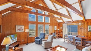 Een ruime woonkamer met houten wanden en een hoge nok, met een groot raam dat uitkijkt op een strand, een eethoek met tafel en stoelen, en een gezellig zithoek met een bank en fauteuils.