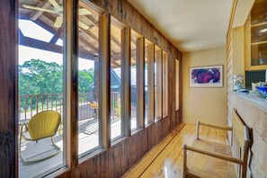Relax in this serene space with a high ceiling, wooden floor, and large window offering a view of the outdoors. A comfortable bench and rocking chair provide seating, while a beautiful artwork adorns the wall.