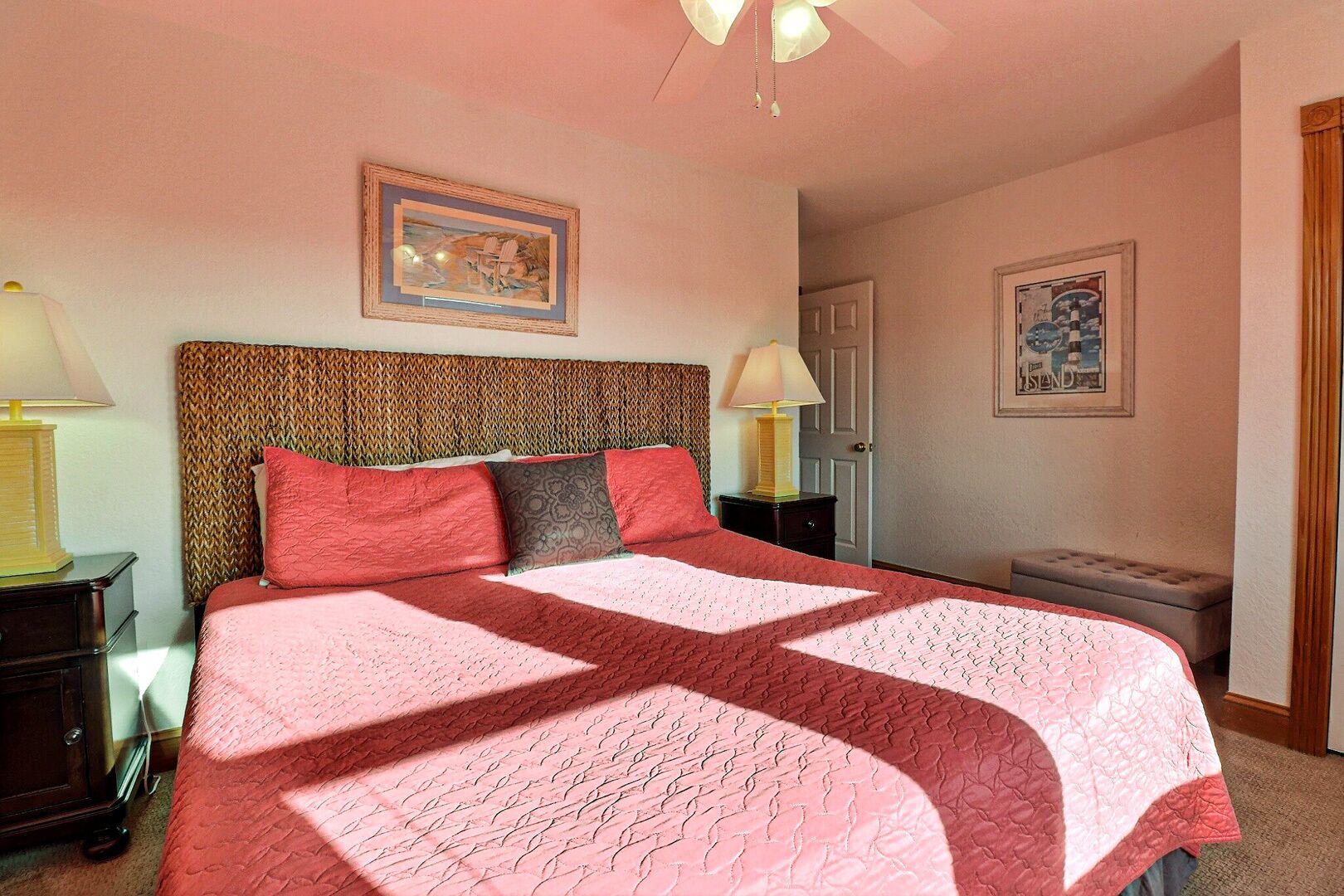 A bedroom with a patterned headboard, a large bed with a pink quilt, two bedside lamps, and a framed picture on the wall.