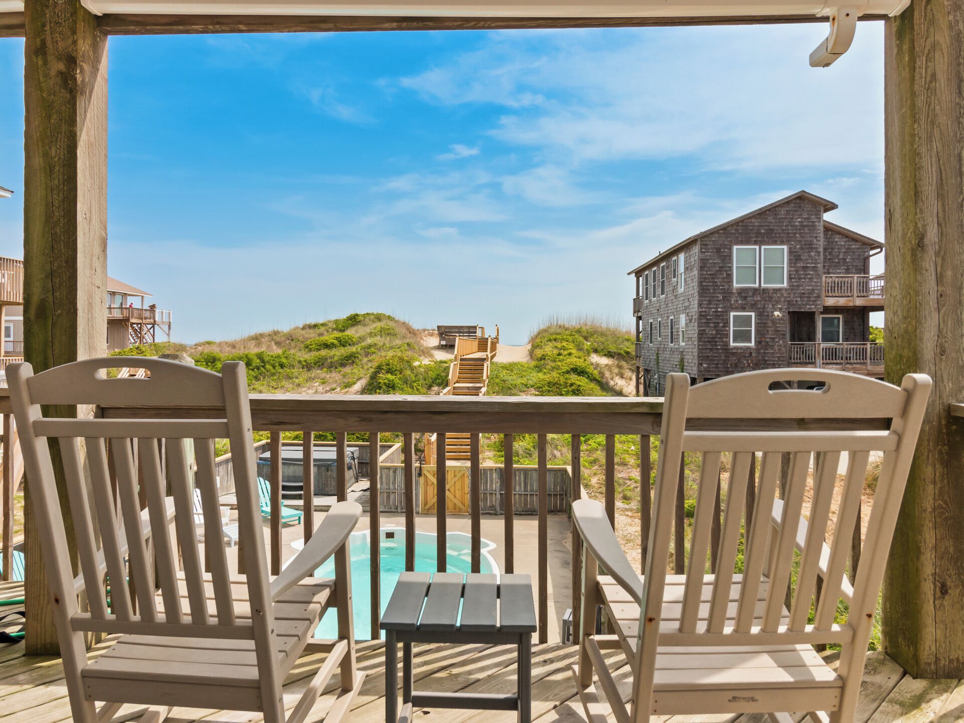 A wooden deck with two Adirondack chairs and a small table, overlooking a swimming pool and a building with a stone facade.