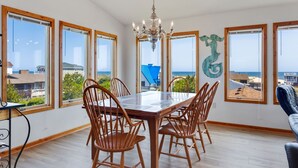 Un espace repas avec une grande table et des chaises en bois, de grandes fenêtres offrant une vue sur l’océan et un lustre.