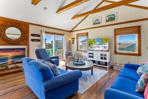 Ein geräumiges Wohnzimmer mit blauen Sofas, einem Couchtisch und einem Fernseher. Die Decke ziert sich mit Holzbalken, ein großes Fenster bietet einen Ausblick und ein Gemälde hängt an der Wand.