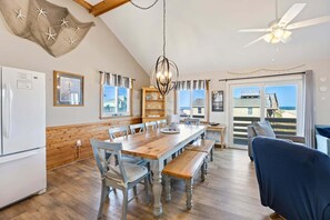 Un coin repas avec une table et des chaises en bois, un lustre et une vue sur la plage par la porte.