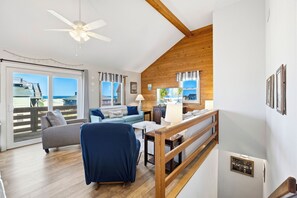 Un salon spacieux avec un plafond en bois, un ventilateur de plafond, une grande fenêtre donnant sur la plage, un canapé, un fauteuil et une petite table.